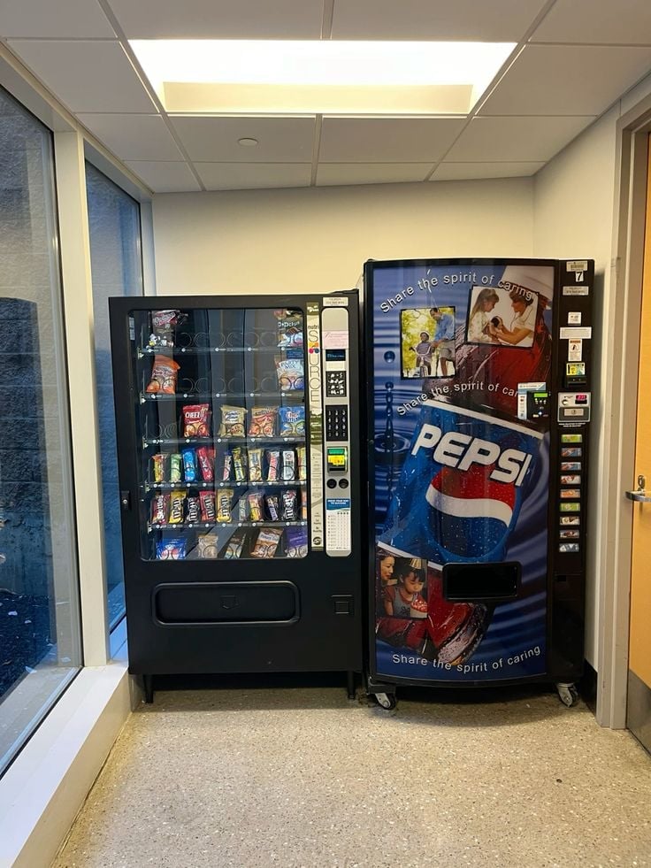 A snack vending machine and Pepsi beverage vending machine side by side in a hallway with beige walls and fluorescent ceiling lights