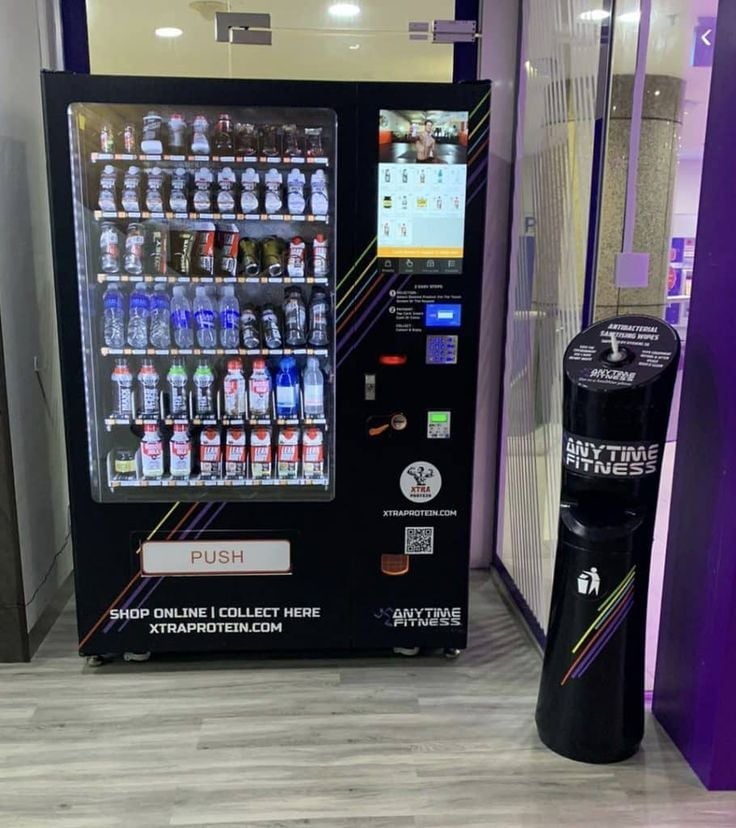 Black vending machine stocked with beverages and supplements in a gym, with a trash bin to the right