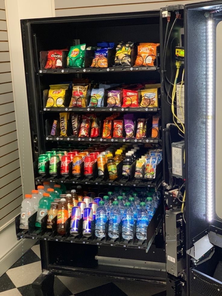 Black vending machine stocked with snacks and beverages including chips, candy, and drinks on multiple shelves