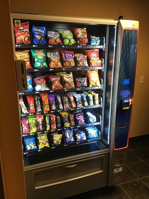 A fully-stocked snack vending machine with colorful chip and candy packages arranged on multiple shelves, featuring a coin/card payment panel on the right side.